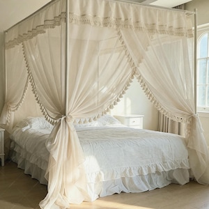 May include: A four-poster bed frame draped with sheer, off-white fabric curtains featuring decorative tassels along the edges. The bed is dressed in a white ruffled bed skirt and white bedding, creating a layered, soft appearance within the frame.