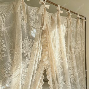 May include: Cream-colored lace curtains with intricate floral and geometric patterns. The curtains are tied with matching ribbons and hang from a wooden rod. The sheer fabric allows light to filter through, creating a soft, romantic ambiance.