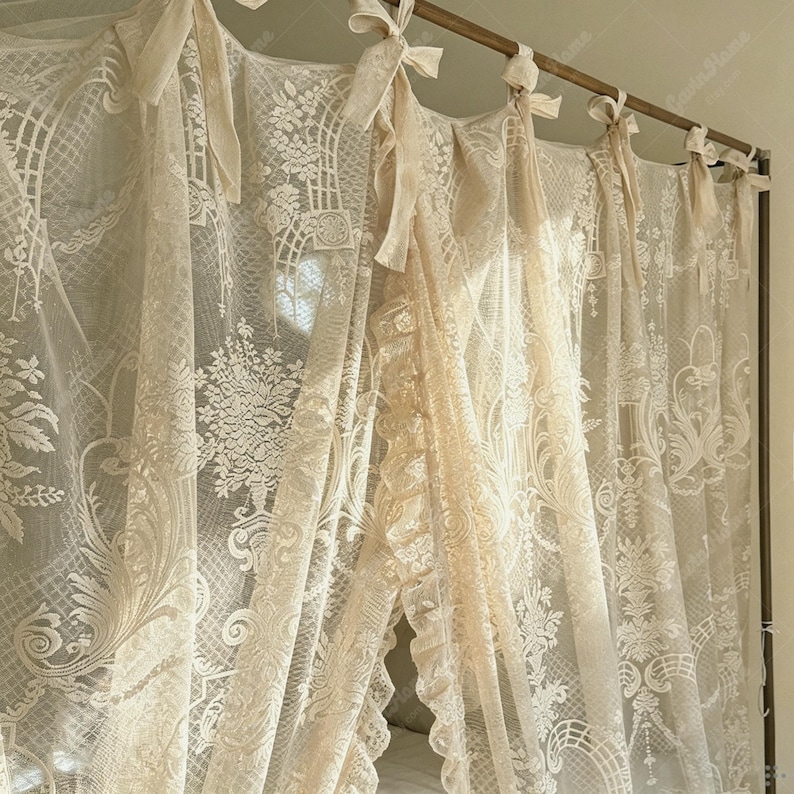 May include: Cream-colored lace curtains with intricate floral and geometric patterns. The curtains are tied with matching ribbons and hang from a wooden rod. The sheer fabric allows light to filter through, creating a soft, romantic ambiance.