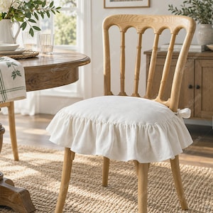 Puede incluir: Una silla de madera clara con un diseño de respaldo de husillos y una funda de asiento de tela blanca. La funda del asiento está adornada con un volante fruncido en los bordes y se sujeta al marco de la silla con pequeñas cintas de tela.
