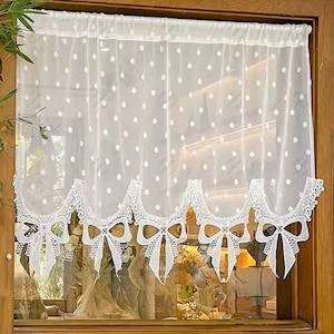 Puede incluir: Cortina blanca de voile con estampado de lunares y un borde inferior festoneado con lazos blancos. La cortina está colgada en un marco de ventana de madera.