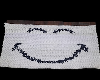 Peut inclure: Un tapis rectangulaire blanc, crocheté, avec un motif de visage souriant. Les yeux et la bouche sont bleu foncé. Le tapis est fait de fil épais et a un aspect texturé. Le fond est noir.