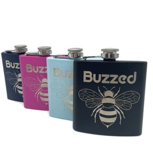 May include: Four stainless steel hip flasks with bee designs and engraved text. The flasks are black, pink, light blue, and dark blue. The text on the flasks reads "Buzz", "Buzzed", and "Buzz".