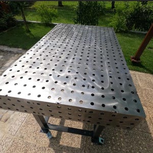 Puede incluir: Una mesa de soldadura metálica rectangular grande con numerosos agujeros espaciados uniformemente. La mesa está soportada por un marco de metal negro con ruedas. La superficie es de color plateado cepillado, reflejando la luz solar. Para uso industrial.