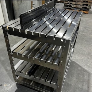 Puede incluir: Una estantería de acero inoxidable con cuatro niveles. Cada nivel tiene listones metálicos horizontales. Una pieza de metal oscuro está en el nivel superior. La unidad está diseñada para uso industrial o comercial.