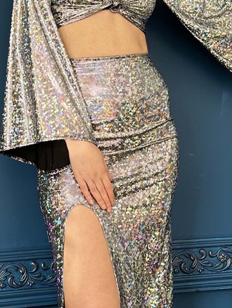 May include: A silver sequined two-piece outfit. The top is a cropped design with long, flared sleeves. The high-waisted skirt features a thigh-high slit. The fabric has a shimmering, iridescent effect, reflecting light.