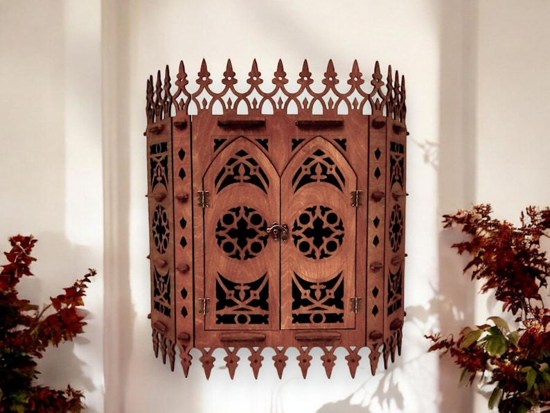 Peut inclure: Une armoire murale en bois de style gothique. L'armoire a deux portes avec des d&eacute;coupes complexes et une arche pointue. L'armoire est peinte en brun fonc&eacute;.