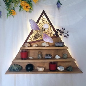 May include: A triangular wooden shelf with three tiers, featuring a cutout of a bee with purple wings and a honeycomb design. The shelf is decorated with crystals, candles, and small jars.