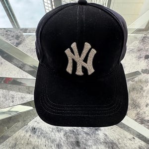 May include: Black baseball cap with a curved peak. The cap has a silver-coloured, rhinestone-studded "NY" logo on the front. The cap is made of a soft, velvet-like material.