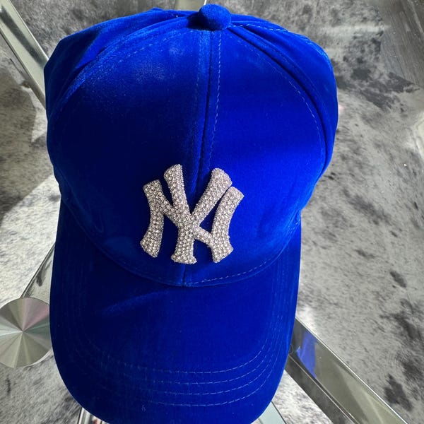 May include: A vibrant royal blue velvet baseball cap featuring a prominent, silver-toned, rhinestone-encrusted NY logo centered on the front panel. The cap has a structured crown, a curved brim with stitched detailing, and a matching fabric button on top.