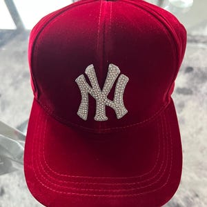 Puede incluir: Una gorra de béisbol de terciopelo rojo intenso con un prominente logotipo NY incrustado con pedrería plateada en el panel frontal. La gorra tiene una corona estructurada, una visera curva con costuras visibles y una textura de tela suave.