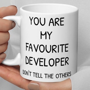 Könnte beinhalten: Weiße Keramiktasse mit schwarzem Text, der lautet: "Du bist mein Lieblingsentwickler. Verrate es nicht weiter."