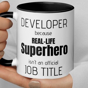 Pode incluir: Caneca de café branca com borda e alça pretas. A caneca tem texto preto que diz "DEVELOPER because REAL-LIFE superhero isn't an official JOB TITLE".