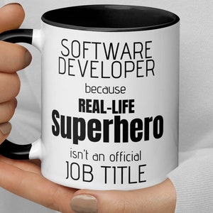 Puede incluir: Taza de café blanca con borde y asa negros. La taza tiene texto negro que dice "Desarrollador de software porque Superhéroe de la vida real no es un título de trabajo oficial."