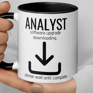 Op de afbeelding: Een witte koffiemok met een zwarte rand en handvat. De mok heeft zwarte tekst die "Analyst software upgrade downloading..." luidt met een zwart pijl-omlaag-pictogram en de tekst "please wait until complete".