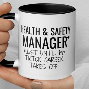 Könnte beinhalten: Eine weiße Keramiktasse mit schwarzem Rand und Henkel. Die Tasse hat einen schwarzen Text mit der Aufschrift "Health & Safety Manager* *Just Until My Tiktok Career Takes Off"