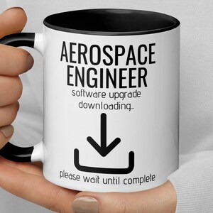 Könnte beinhalten: Eine weiße Kaffeetasse mit schwarzem Rand und Henkel. Die Tasse hat einen schwarzen Text, der "Aerospace Engineer software upgrade downloading." lautet. Unter dem Text befindet sich ein schwarzes Abwärtspfeilsymbol. Der Text "please wait until complete" befindet sich unter dem Symbol.