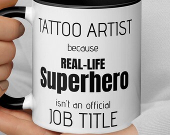 Könnte beinhalten: Eine weiße Keramiktasse mit schwarzem Rand und Henkel. Die Tasse hat einen schwarzen Text mit der Aufschrift "Tattoo Artist because Real-Life Superhero isn't an official Job Title."