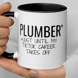 Puede incluir: Taza de café blanca con borde y asa negros. La taza tiene texto negro que dice "Plumber* *Just Until My Tiktok Career Takes Off".