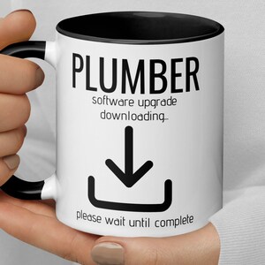 Puede incluir: Taza de café blanca con borde y asa negros. La taza tiene texto negro que dice "Plumber software upgrade downloading." con un icono de flecha negra hacia abajo y el texto "please wait until complete".
