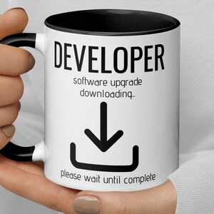 Op de afbeelding: Witte keramische mok met een zwarte rand en handvat. De mok heeft zwarte tekst die "DEVELOPER software upgrade downloading_" luidt met een zwart pijl-omlaag-pictogram en de tekst "please wait until complete".