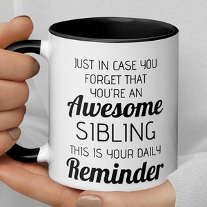 Op de afbeelding: Een witte koffiemok met een zwarte handgreep en een zwarte rand. De mok heeft zwarte tekst die luidt "Just in case you forget that you're an Awesome Sibling. This is your daily Reminder."