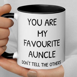 Può includere: Tazza in ceramica bianca con bordo e manico neri. La tazza ha un testo nero che recita "You are my favourite auncle. Don't tell the others."