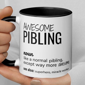 Può includere: Una tazza da caffè bianca con un bordo e un manico neri. La tazza ha un testo nero che recita "AWESOME PIBLING noun like a normal pibling, except way more awesome see also: superhero, miracle worker"