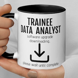 Op de afbeelding: Een witte koffiemok met een zwarte rand en handvat. De mok heeft zwarte tekst die "Trainee Data Analyst" luidt met een downloadpijl en de tekst "software-upgrade wordt gedownload. wacht alstublieft tot het proces is voltooid".