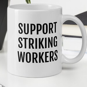Op de afbeelding: Een witte keramische mok met zwarte tekst die "SUPPORT STRIKING WORKERS" luidt.