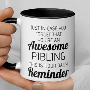 Può includere: Una tazza da caffè bianca con un bordo e un manico neri. La tazza ha un testo nero che recita "Just in case you forget that you're an Awesome Sibling. This is your daily reminder."