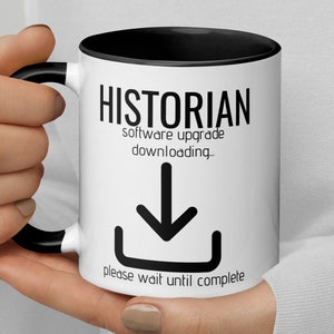 May include: White coffee mug with a black rim and handle. The mug has black text that reads "Historian software upgrade downloading_" with a black downward arrow and the text "please wait until complete".
