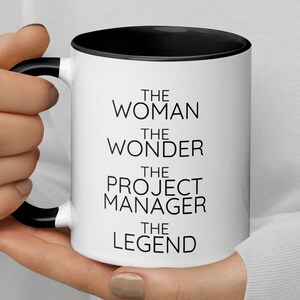 May include: White ceramic mug with a black rim and handle. The mug has black text that reads "The Woman The Wonder The Project Manager The Legend".