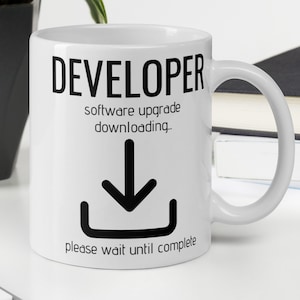 Op de afbeelding: Een witte keramische koffiemok met een zwarte afbeelding waarop staat "DEVELOPER software-upgrade wordt gedownload..." met een pijl naar beneden en een vak eronder. De tekst onder de pijl luidt "wacht alstublieft tot de voltooiing."