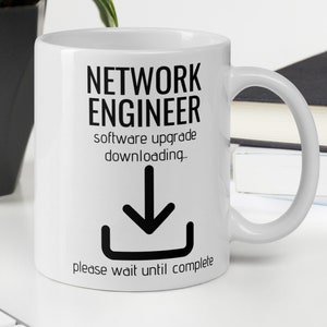 Pode incluir: Caneca de café de cerâmica branca com um design preto que diz "Network Engineer software upgrade downloading... please wait until complete" com um ícone de seta preta para baixo.