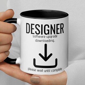 Könnte beinhalten: Weiße Keramiktasse mit schwarzem Rand und Henkel. Die Tasse hat schwarzen Text, der "DESIGNER software upgrade downloading." lautet. Unter dem Text befindet sich ein schwarzes Abwärtspfeilsymbol. Der Text "please wait until complete" befindet sich unter dem Symbol.