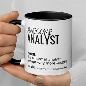 Op de afbeelding: Een witte koffiemok met een zwarte rand en handvat. De mok heeft zwarte tekst met de tekst "AWESOME ANALYST noun like a normal analyst, except way more awesome see also: superhero, miracle worker"