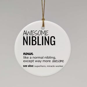 Op de afbeelding: Witte keramische ornament met zwarte tekst die luidt: "AWESOME NIBLING zelfstandig naamwoord zoals een normale neef of nicht, behalve veel meer awesome zie ook: superheld, wonderdoener"