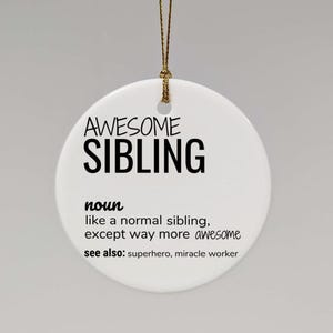 Op de afbeelding: Witte keramische ornament met zwarte tekst die "AWESOME SIBLING noun like a normal sibling, except way more awesome see also: superhero, miracle worker" luidt.
