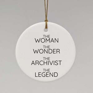 Könnte beinhalten: Ein weißer, runder Keramikschmuckanhänger mit goldener Schnur. Der Anhänger trägt den schwarzen Text "THE WOMAN THE WONDER THE ARCHIVIST THE LEGEND". Ein schlichtes, elegantes Design.