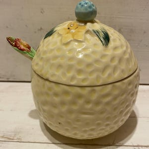 May include: A yellow ceramic honey pot with a lid and a small spoon. The pot is decorated with a flower and a blue knob on the lid.