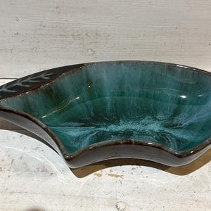 May include: A teal ceramic serving dish with an unusual, asymmetrical form. The interior has a glossy finish, fading to a darker brown rim. A leaf design is etched on one end. Suitable for serving snacks or as a decorative item. Measures approximately 30cm in length.