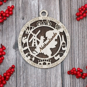 May include: A round wooden ornament with a map of the Netherlands, featuring cut-out designs of a windmill, bicycle, and other Dutch icons. The word "Netherlands" is carved into the wood. Red berries are arranged around the ornament.