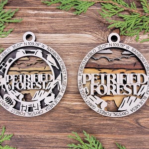 May include: Two wooden ornaments with a laser-cut design of the Petrified Forest National Park in Arizona. The ornaments are round and feature the park's name, location, and coordinates. The design includes a stylized image of a petrified log, a bird, and a mountain.