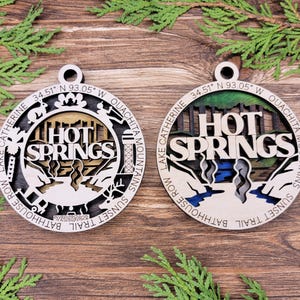 May include: Two wooden ornaments with a laser-cut design of the Hot Springs, Arkansas city logo. The text on the ornaments reads "Hot Springs" with the coordinates "34.51°N 93.05°W" and "Ouachita Mountains". One ornament has a blue and green color scheme, while the other has a brown and gold color scheme.