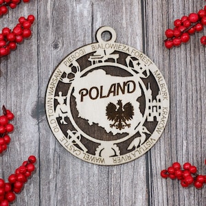 May include: Wooden ornament featuring a map of Poland with the word "Poland" in the center. The ornament is circular with the names of Polish cities and landmarks around the edge. The ornament is surrounded by red berries.