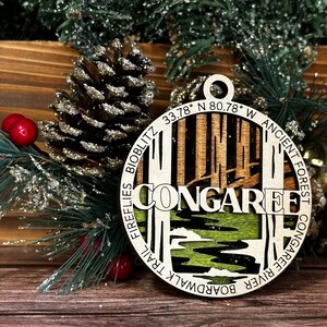 May include: A wooden ornament with a laser-cut design depicting the Congaree National Park in South Carolina. The ornament features the park's name, coordinates, and a description of the park's features, including the boardwalk trail, fireflies, and the ancient forest.