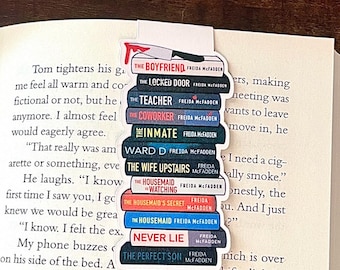 May include: A stack of Freida McFadden book-themed bookmarks, each with a book title. The bookmarks are placed on an open book with text. The books are stacked on top of each other.