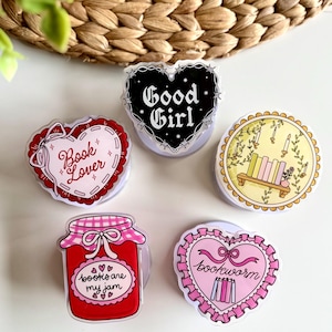 May include: A collection of five colorful phone grips with various designs. One features a black heart with "Good Girl" text, another a red heart with "Book Lover", and a third with "Books are my jam". The other two have a book theme.