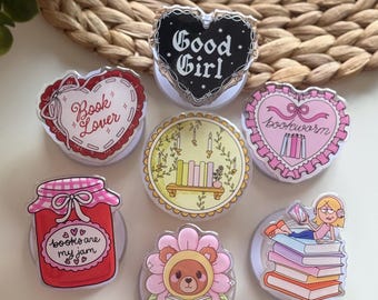 May include: A collection of seven circular phone grips featuring various designs: a black heart with 'Good Girl', a red heart with 'Book Lover', a pink heart with 'bookworm', a jar with 'books are my jam', a shelf of books, a bear in a flower, and a person reading on a stack of books.